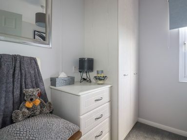 A corner of a room with a dresser topped with a lamp tissue box and candle next to a wardrobe and a chair with a cushion and teddy bear at Lowen Lodge St Merryn near Padstow