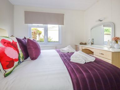 A bedroom with a bed covered with purple bed runner and pillows beside a dresser with a mirror at Lowen Lodge in St Merryn near Padstow