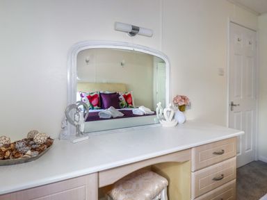 A bedroom vanity desk with a large mirror reflecting a bed with floral and purple pillows and towels at Lowen Lodge in St Merryn near Padstow