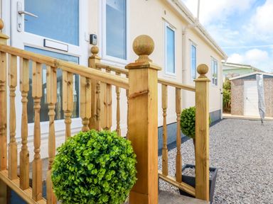 A wooden railing with decorative posts next to a white door and windows with potted green plants on a gravel pathway at Lowen Lodge in St Merryn near Padstow