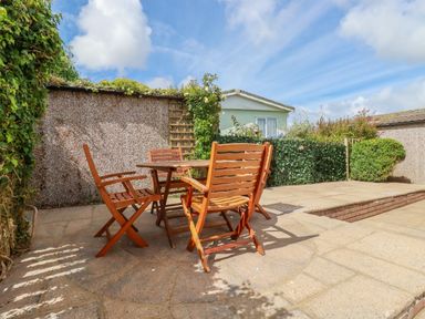 An outdoor patio area with a wooden table and chairs surrounded by hedges and a building in the background at Lowen Lodge in St Merryn near Padstow