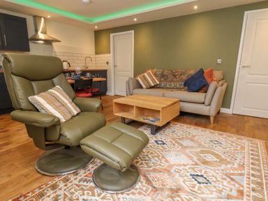 A living room with an olive green armchair and ottoman a wooden coffee table a sofa with cushions a patterned rug and a small kitchen in the background at Picadore Two in Scarborough
