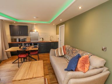A room with a sofa with pillows a wooden coffee table a dining table set with plates glasses and cutlery and a kitchen area with cabinets appliances and a sink at Picadore Two in Scarborough