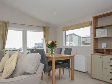 A dining area with a wooden table and four chairs next to a gray sofa with yellow pillows and a cabinet with shelves at Lodge 74 at Riviera Bay Coastal Retreat in Brixham