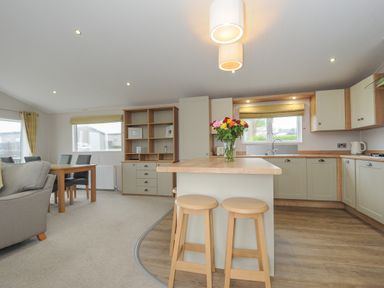 A kitchen with an island with stools and flowers a dining table and chairs and a shelving unit at Lodge 74 at Riviera Bay Coastal Retreat in Brixham