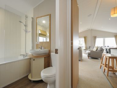 A bathroom with a bathtub shower combination a sink and a toilet next to an open door leading to a living area with chairs stools and windows at Lodge 74 at Riviera Bay Coastal Retreat in Brixham