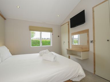 A bedroom with a bed, rolled towels on the bed, a window, a wall-mounted television, and a wall-mounted shelf with a mirror at Lodge 74 at Riviera Bay Coastal Retreat in Brixham