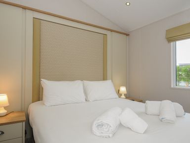 A bedroom with a double bed white pillows rolled towels on bed bedside tables with lamps and a window at Lodge 74 at Riviera Bay Coastal Retreat Brixham