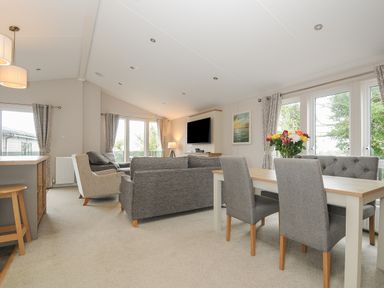 A living room with grey sofas a dining table with chairs and flowers on the table at Lodge 59 at Riviera Bay Coastal Retreat in Brixham