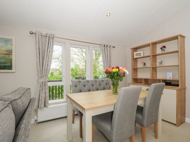 A dining area with a wooden table and grey chairs next to windows and a wooden shelving unit at Lodge 59 at Riviera Bay Coastal Retreat in Brixham