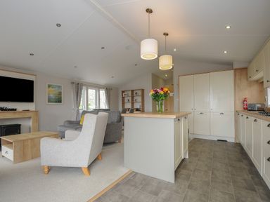 A kitchen with a central island and pendant lights next to a living room with armchairs a coffee table and a wall-mounted television at Lodge 59 at Riviera Bay Coastal Retreat in Brixham