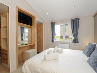 A bedroom with a double bed with three rolled towels on it a window with blue patterned curtains a wall-mounted tv and a dressing table with a stool at Lodge 59 at Riviera Bay Coastal Retreat in Brixham