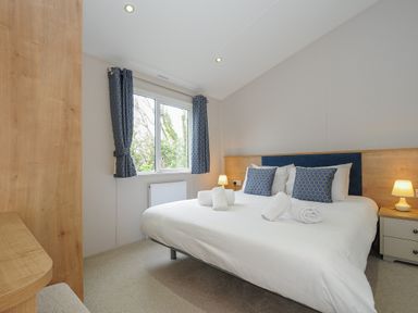 A bedroom with a double bed with pillows and rolled towels beside a window with blue curtains at Lodge 59 at Riviera Bay Coastal Retreat in Brixham