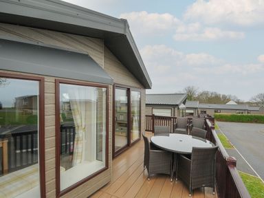 An outdoor balcony with a round glass table and wicker chairs attached to a wooden cabin at Lodge 69 at Riviera Bay Coastal Retreat in Brixham