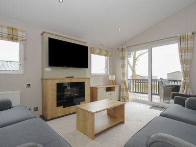 A living room with gray sofas a wooden coffee table a mounted TV above a fireplace and sliding glass doors at Lodge 69 at Riviera Bay Coastal Retreat in Brixham