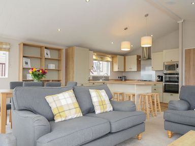 A living room with gray sofas and yellow plaid cushions next to a kitchen with wooden stools and cabinets at Lodge 69 at Riviera Bay Coastal Retreat in Brixham