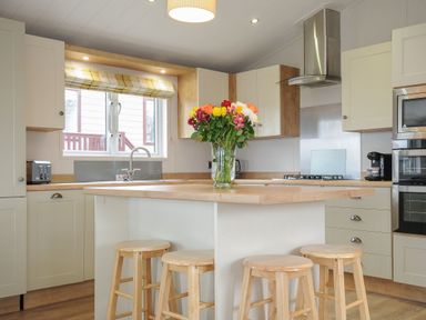 A kitchen with wooden stools around an island and a vase of flowers on the countertop at Lodge 69 at Riviera Bay Coastal Retreat in Brixham