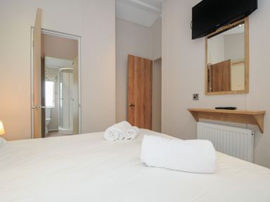 A bedroom with a bed and rolled towels a mirror with a wooden frame below a mounted TV and an open door showing a bathroom at Lodge 69 at Riviera Bay Coastal Retreat in Brixham