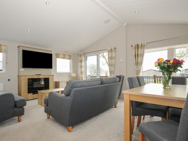 A living room with grey sofas a dining table with flowers and a TV above a fireplace at Lodge 68 at Riviera Bay Coastal Retreat in Brixham