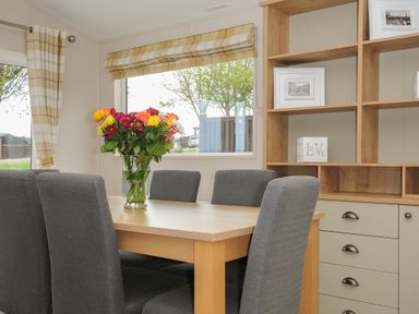 A dining area with a wooden table six gray chairs a vase of flowers and shelving at Lodge 68 at Riviera Bay Coastal Retreat Brixham
