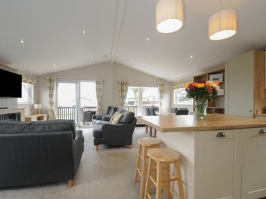 An open plan living area with grey sofas a wall mounted television a wooden kitchen island with stools and a dining table at Lodge 68 at Riviera Bay Coastal Retreat in Brixham
