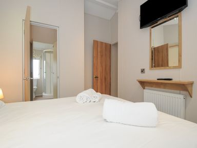 A bedroom with a bed and rolled towels a wall-mounted TV a mirror and an open door leading to a bathroom at Lodge 68 at Riviera Bay Coastal Retreat in Brixham