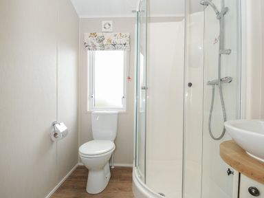 A bathroom with a toilet, a window with floral blind, a glass shower enclosure, and a sink on a wooden countertop at Lodge 68 at Riviera Bay Coastal Retreat Brixham