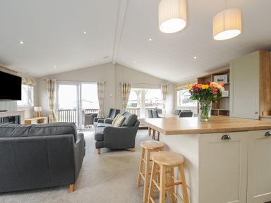 A living area with grey sofas wooden stools a kitchen island with flowers and a dining table with chairs at Lodge 70 at Riviera Bay Coastal Retreat in Brixham