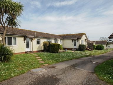 A row of single-story houses with tiled roofs and green lawns along a paved pathway at Chalet 7 at Landscove Holiday Park in Brixham