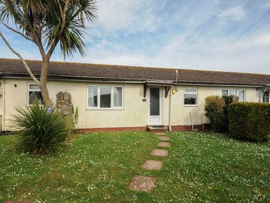 A single-story house with a tiled roof a white door and windows a grassy lawn with stepping stones and trees and bushes at Chalet 7 at Landscove Holiday Park Brixham