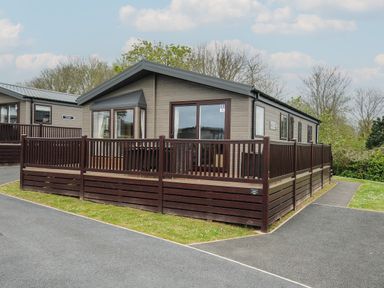 A single story cabin with a wooden deck and railing surrounded by pavement and grass at Lodge 72 at Riviera Bay Coastal Retreat in Brixham