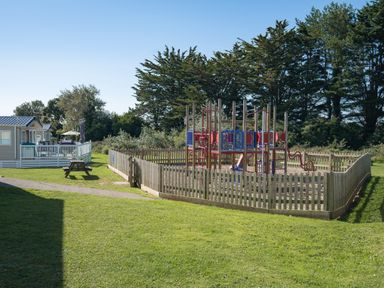 A playground with climbing equipment surrounded by a wooden fence next to a picnic bench and nearby holiday park buildings at Chalet 31 at Landscove Holiday Park in Brixham
