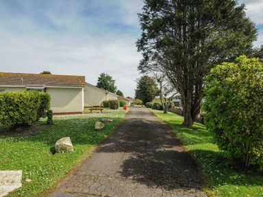 A paved pathway flanked by grass with bushes and trees next to low single-story buildings at Chalet 15 at Landscove Holiday Park in Brixham