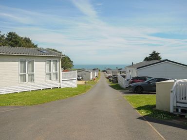 A paved road lined with white holiday chalets and parked cars leading to the sea at Chalet 15 at Landscove Holiday Park in Brixham