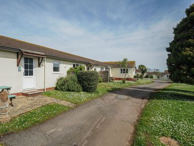 A row of single-story holiday chalets with paved paths and grassy areas with bushes and trees at Chalet 34 at Landscove Holiday Park in Brixham