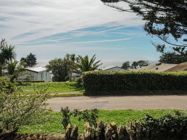 A view of a grassy area with trees and bushes overlooking several buildings and the sea at Chalet 34 at Landscove Holiday Park in Brixham