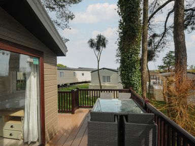 An outdoor deck with a glass table and chairs next to a house with other mobile homes and trees in the background at Lodge 15 at Landscove Holiday Park in Brixham