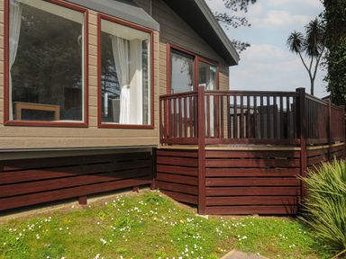 An exterior view of a wooden lodge with a deck and a grassy area with small white flowers at Lodge 15 at Landscove Holiday Park in Brixham
