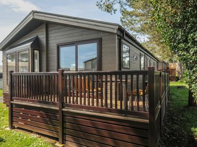 A holiday lodge with a wooden deck and outdoor chairs surrounded by grass and trees at Lodge 9 at Landscove Holiday Park in Brixham
