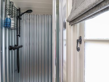 A shower area with corrugated metal walls a black showerhead and a window with a gray Roman shade at La'al Ling in Penrith