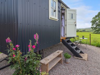 An outdoor area with a black corrugated metal tiny house with white door open showing a robe a wooden bench pink flowers a potted plant black metal stairs and a small black table and chairs set on gravel at La'al Ling in Penrith