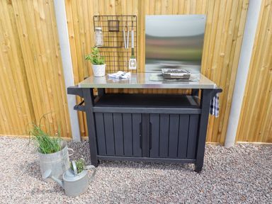 An outdoor barbecue station with a metal countertop and a black storage cabinet against a wooden fence at La'al Ling in Penrith