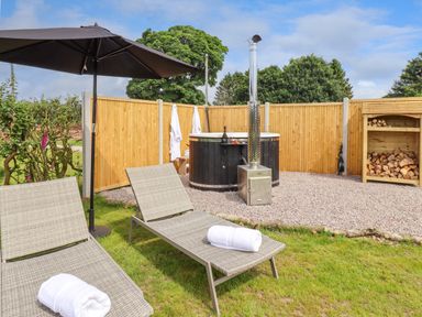 An outdoor area with two lounge chairs and a large black umbrella near a hot tub with a chimney and a wooden fence at La'al Ling in Penrith