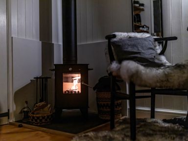 A small wood-burning stove with fire inside and a chair with a fur throw in a corner of a room at La'al Ling in Penrith