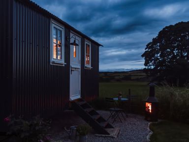 An exterior view of a small building with lit windows a door with stairs a table with two chairs and a lit outdoor fireplace at La'al Ling Penrith