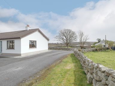 A white house with a brown roof next to a paved driveway and stone walls in a rural area at Rossaveel Upper in Rossaveel County Galway