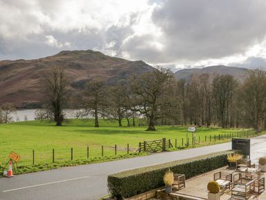 A road with a roadside patio and chairs overlooking green fields with trees and hills in the background at Ullswater View Apartment Ullswater