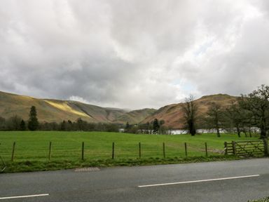 A road with a wooden fence and green field with hills and trees in the background at Ullswater View Apartment in Ullswater