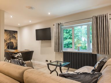 A living room with a beige sofa a glass coffee table a wall mounted TV a dining table with chairs and a window with curtains at Ullswater View Apartment in Ullswater