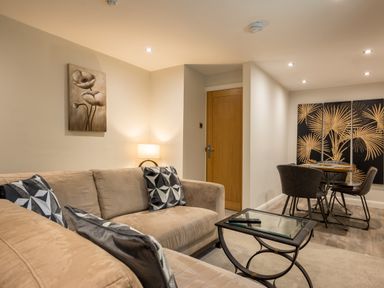A living room with beige sofas and patterned cushions a lamp a glass coffee table and a dining table with chairs and artwork on the walls at Ullswater View Apartment Ullswater